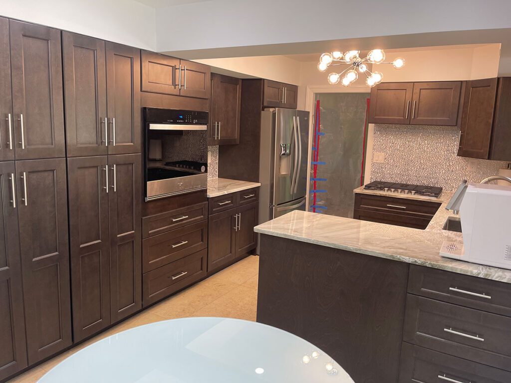 Modern kitchen with dark wooden cabinets, stainless steel appliances including a stove, oven, and fridge, marble countertops, and a round light fixture. A round glass table is in the foreground.