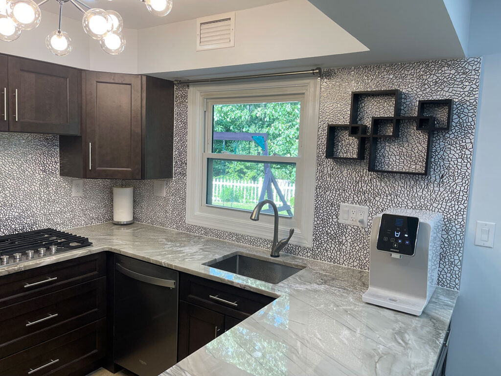 A modern kitchen with dark cabinets, marble countertops, mosaic tile backsplash, a single window, a stainless steel sink, a mounted black geometric shelf, and a countertop water dispenser.