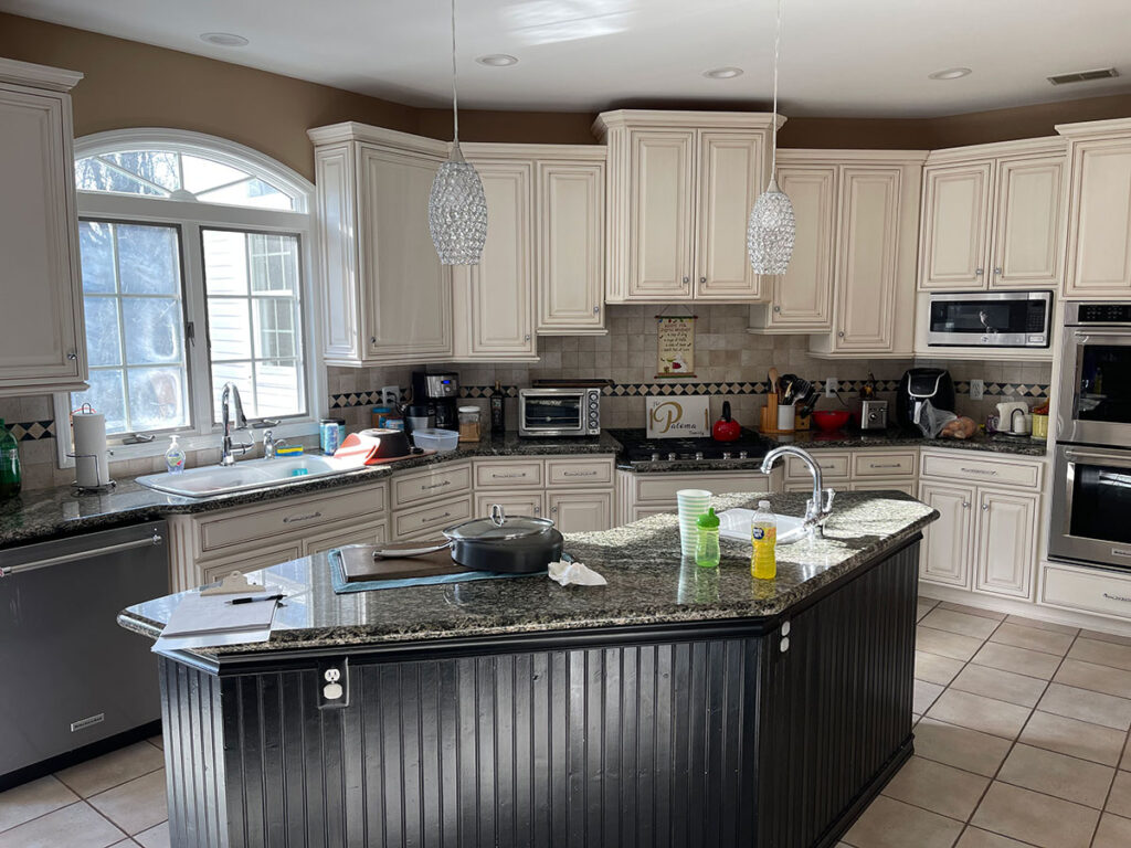 A modern kitchen with white cabinets, stainless steel appliances, a central island with a sink, and various items on the countertops. Two pendant lights hang above the island. Sunlight filters in through a window.