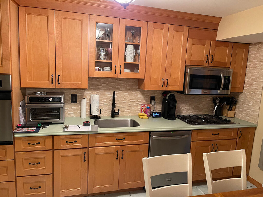 A kitchen with wooden cabinets, stainless steel appliances, a countertop with various items, a sink, and a tiled backsplash. A dining table and chairs are in the foreground.